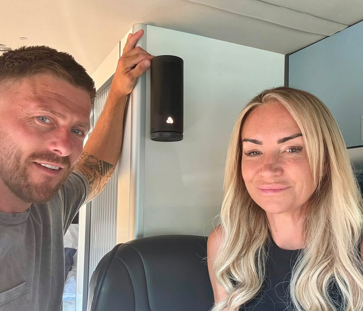 A man and a woman inside a vehicle or camper, with the man pointing to a black PINE Beat cylindrical speaker mounted on a light gray cabinet.