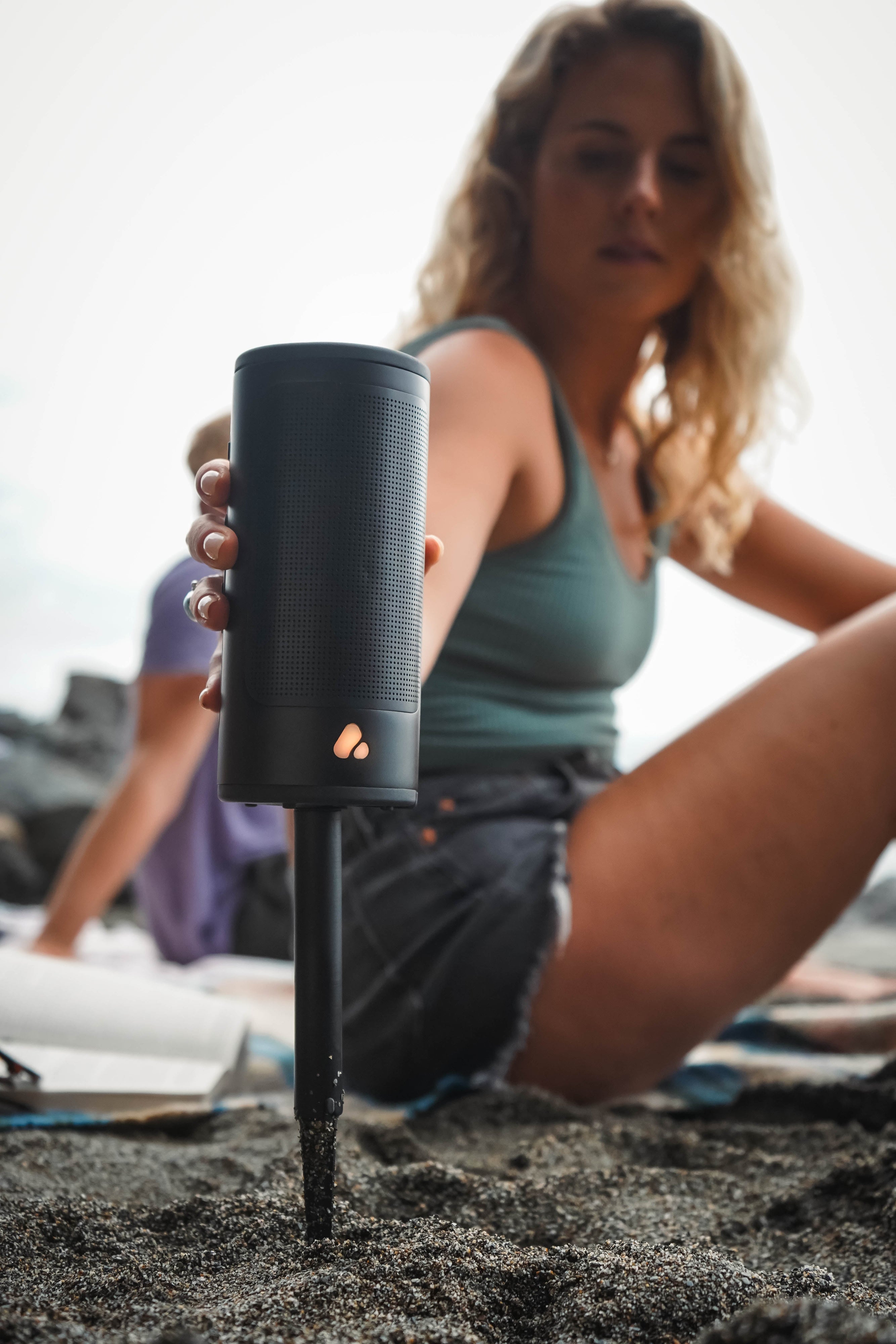 Woman sitting on beach holding a black portable speaker with a stake attachment underneath, she is staking it on the sand.