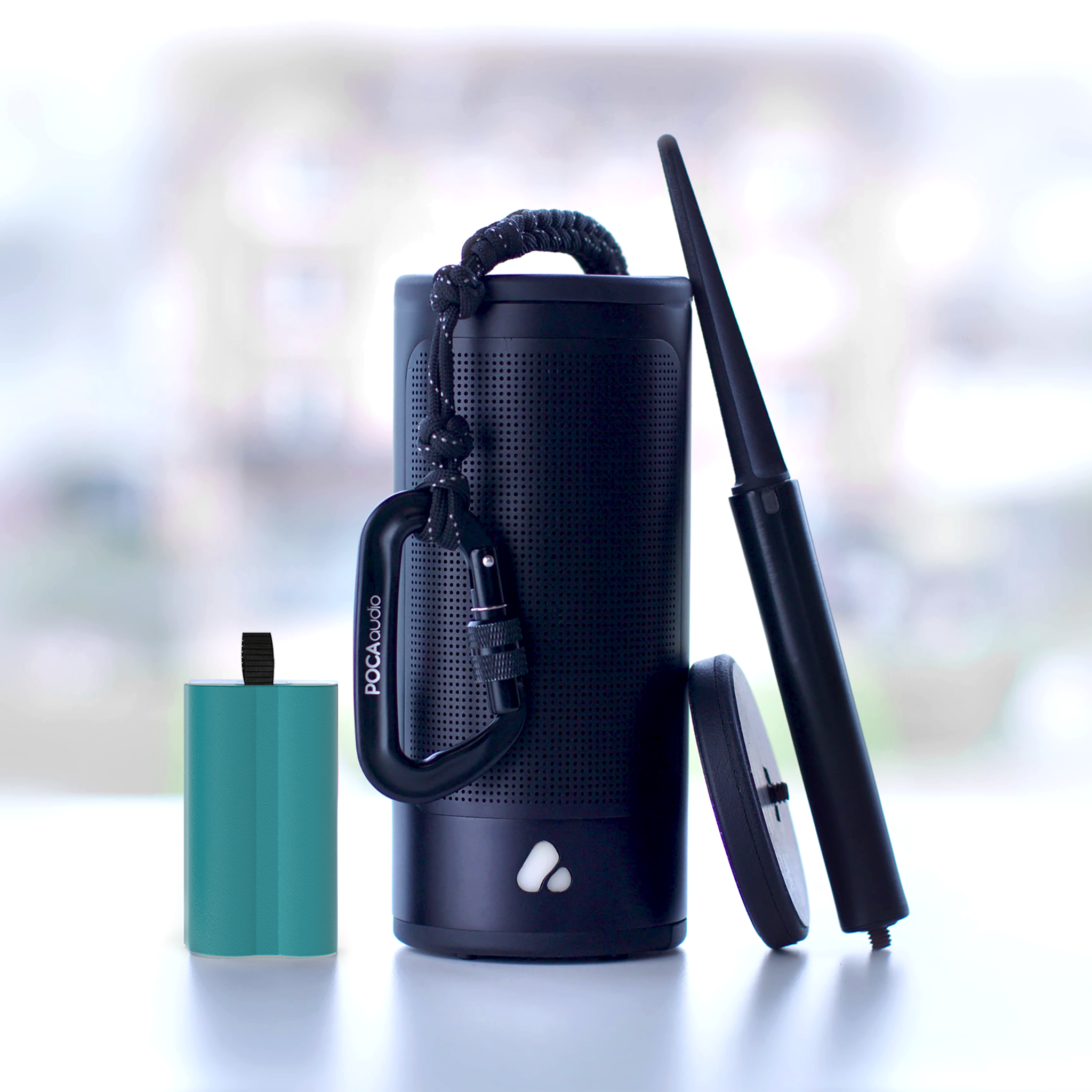 Cylindrical black speaker surrounded by accessories including a blue battery a black stake, a disc-like magnetic base and a rope with a POCA audio branded carabiner.