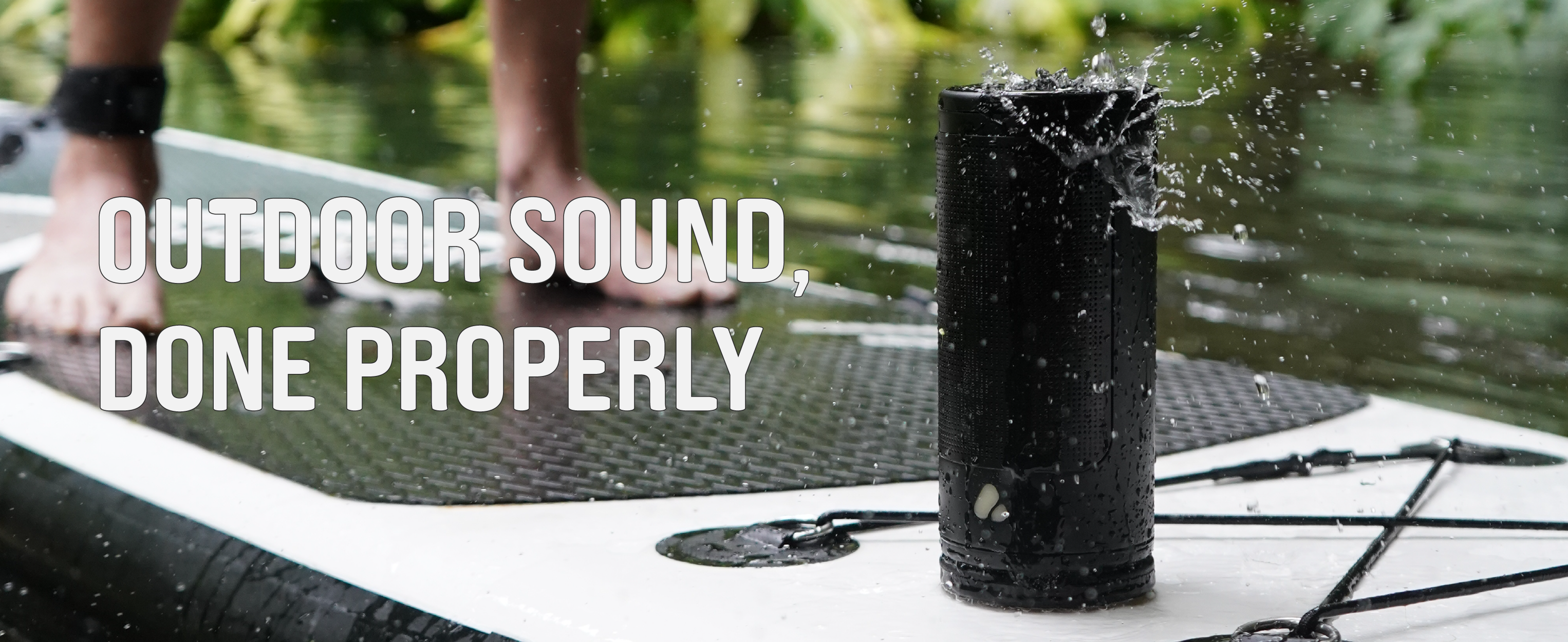 A black outdoor speaker standing on a paddleboard on water, with a person's bare feet visible in the background and large white text reading 'OUTDOOR SOUND, DONE PROPERLY'.