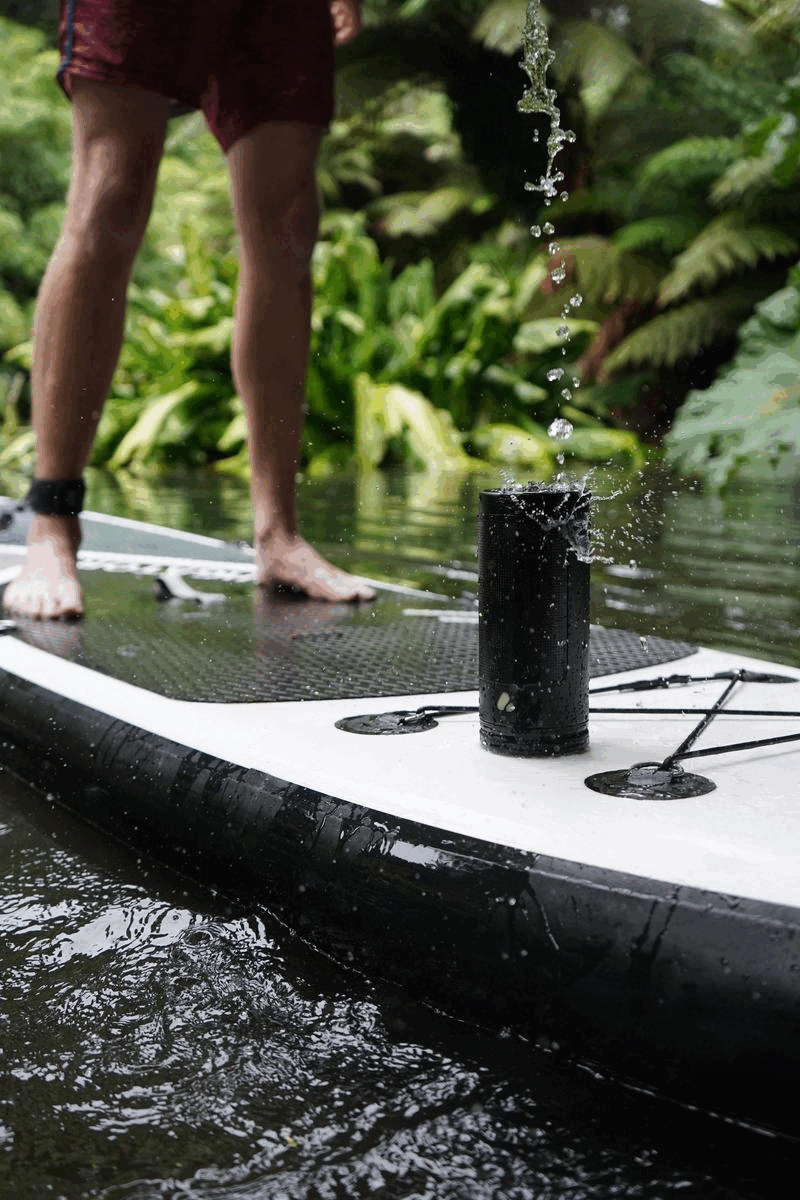 Cylindrical speaker attached to paddleboard on river and forest background, paddle splashing water on top of speaker.