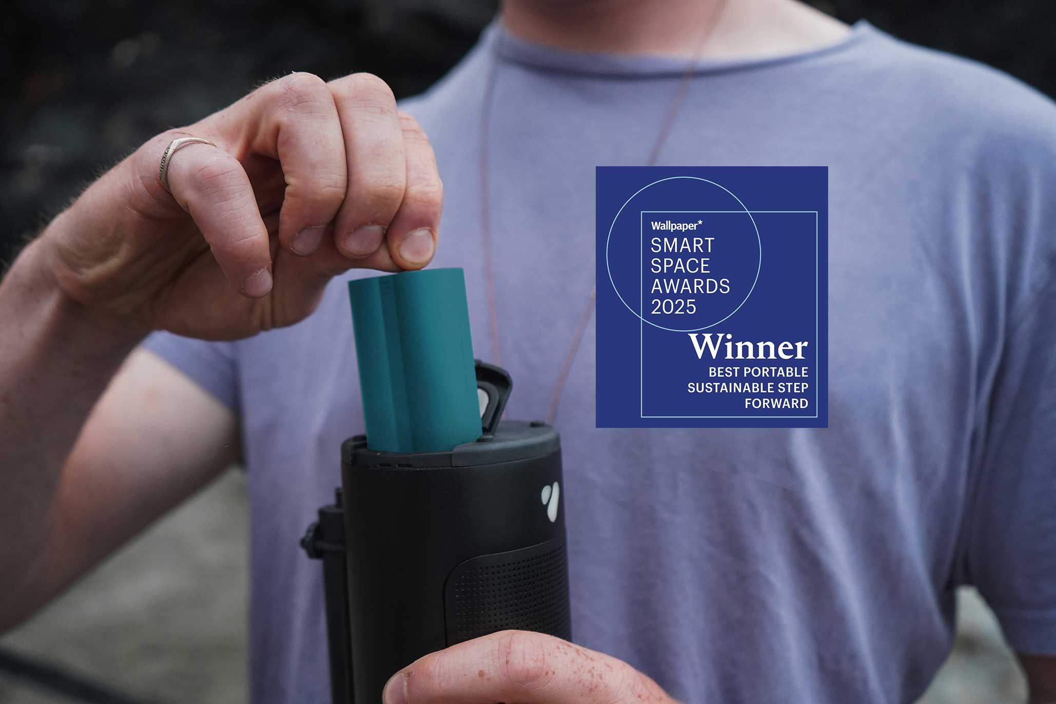 Person pulling a teal battery out of a black portable speaker with a 'Wallpaper Smart Space Awards 2025 Winner' badge on a blurred background