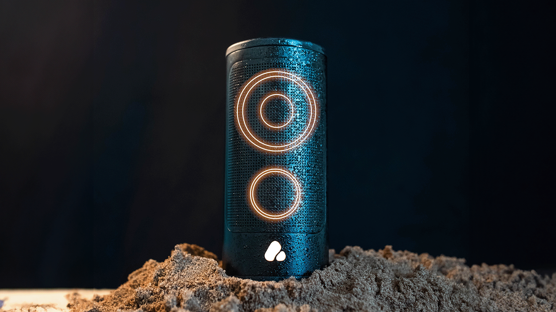 Cylindrical speaker on sand with black background and glowing circles indicating tweeter and woofer placement