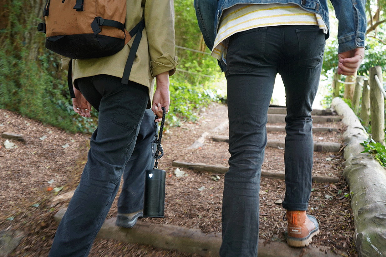two people walking up some gravel steps, between them is a portable speaker being held by a rope accessory and lights are on.