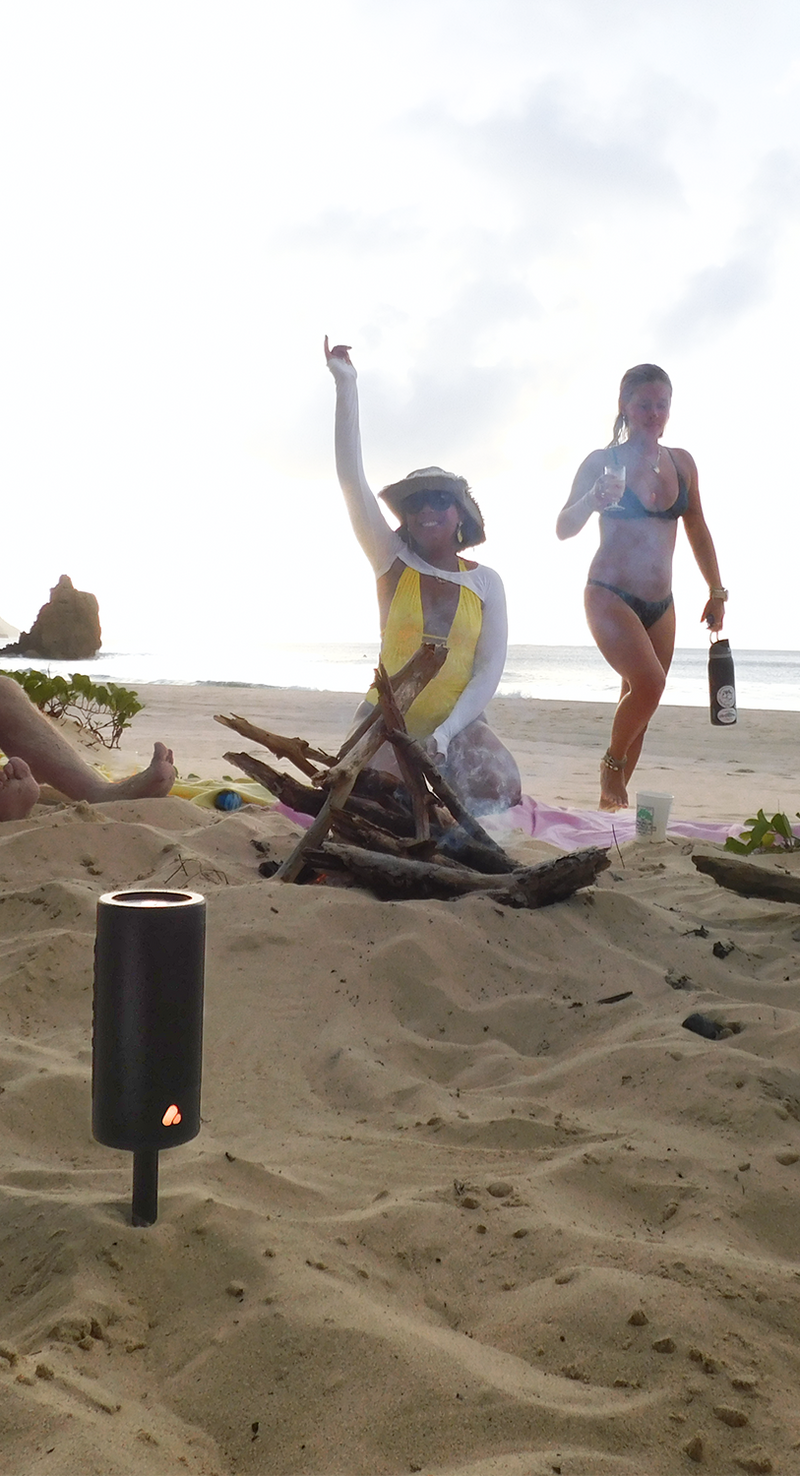 People on a beach setting up a fire pit with sand and natural elements listening to music from speaker staked on sand.