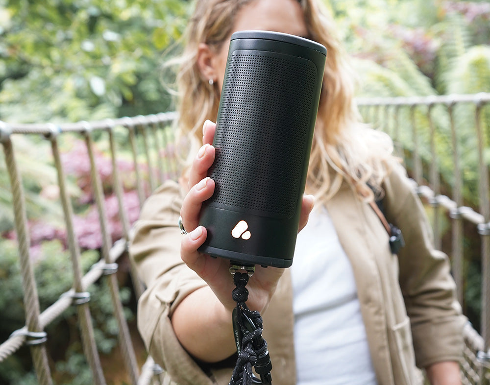 Woman holding a black cylindrical POCA audio speaker, outdoors.