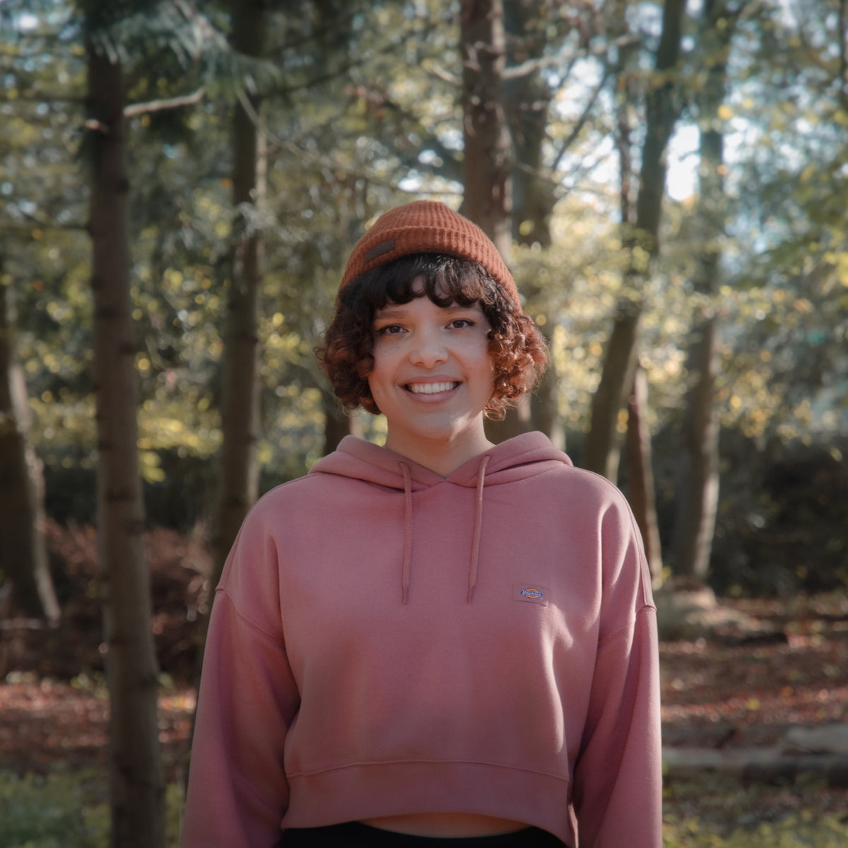portrait of smiling woman in front of a forest wearing a brown beanie and thick mauve pink hoodie 